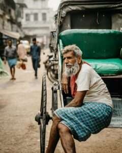 An elderly rickshaw puller with a gray beard and a contemplative expression sits on the edge of his rickshaw in a busy urban street. He wears a white shirt, a red towel draped around his neck, and a blue checkered lungi. The vibrant green seat of the rickshaw contrasts with the muted tones of the bustling street in the background, where pedestrians and vehicles move through the hazy atmosphere, capturing a moment of stillness amidst city life