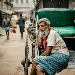 An elderly rickshaw puller with a gray beard and a contemplative expression sits on the edge of his rickshaw in a busy urban street. He wears a white shirt, a red towel draped around his neck, and a blue checkered lungi. The vibrant green seat of the rickshaw contrasts with the muted tones of the bustling street in the background, where pedestrians and vehicles move through the hazy atmosphere, capturing a moment of stillness amidst city life