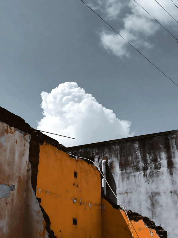 This image portrays a stark contrast between decay and beauty- a crumbling yellow wall, with its peeling paint and exposed structure, tells a story of time and neglect. Above it, the crisp white clouds stand out against the muted sky, adding a sense of calm and hope.