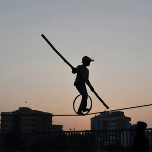 This photograph captures the silhouette of a young tightrope walker balancing skillfully on a rope with a unicycle, holding a balancing pole. The backdrop of the setting sun and urban skyline enhances the contrast, emphasizing the figure's courage and determination.