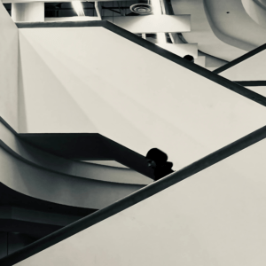 This black-and-white photograph presents a striking interplay of geometric shapes and abstract design in urban architecture. The minimalist composition, with its stark lines and contrasting light and shadow, creates a sense of depth with the rush of movement on figures on the staircase.