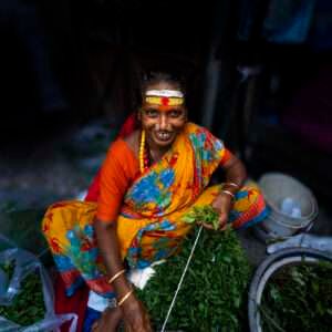 This captivating portrait features a woman dressed in a vibrant saree with bold colors and floral patterns. She is seated amidst fresh greens sorting malas for sale. The background is blurred, directing focus to her vibrant dress, adornments and wide smile.