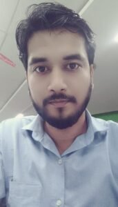 Manni, a young man with short black hair and a trimmed beard, is wearing a light blue shirt and looking into the camera indoors, with a blurred office background. 