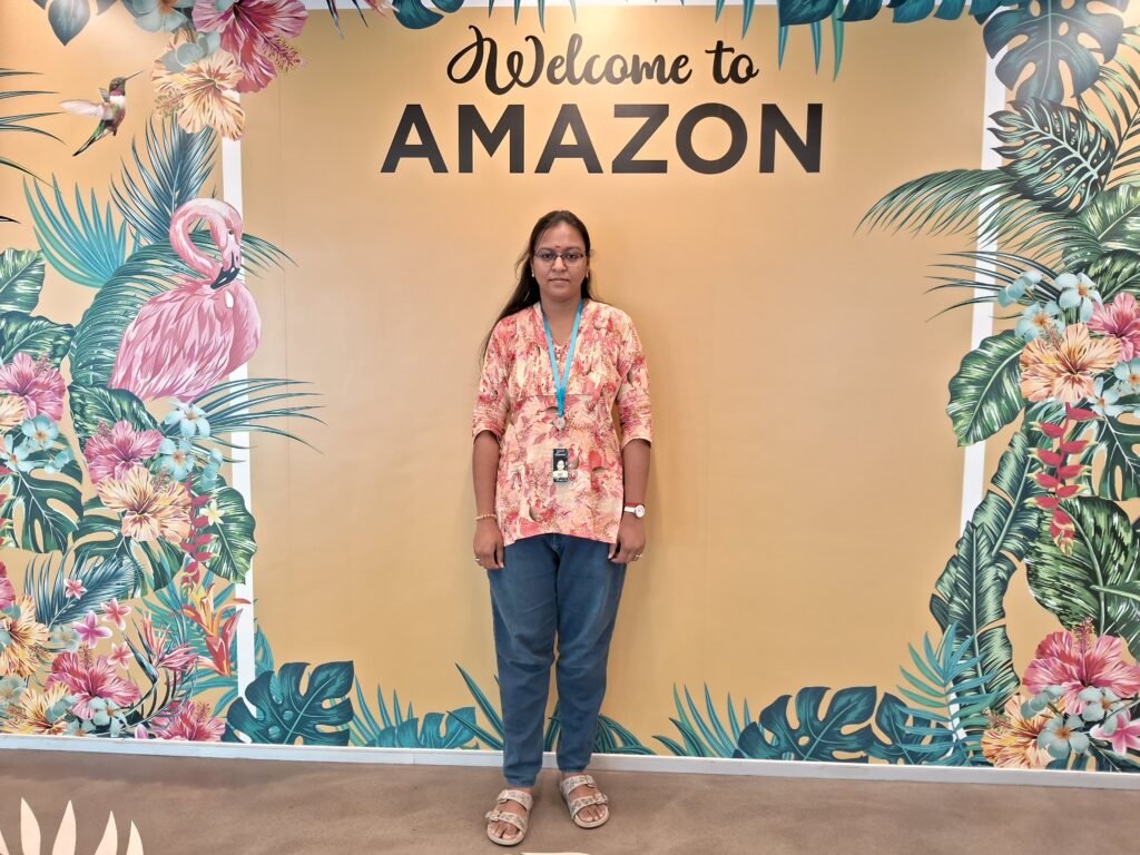 A woman wearing a floral top and ID badge stands in front of a vibrant "Welcome to Amazon" wall decorated with tropical leaves and pink flamingos.