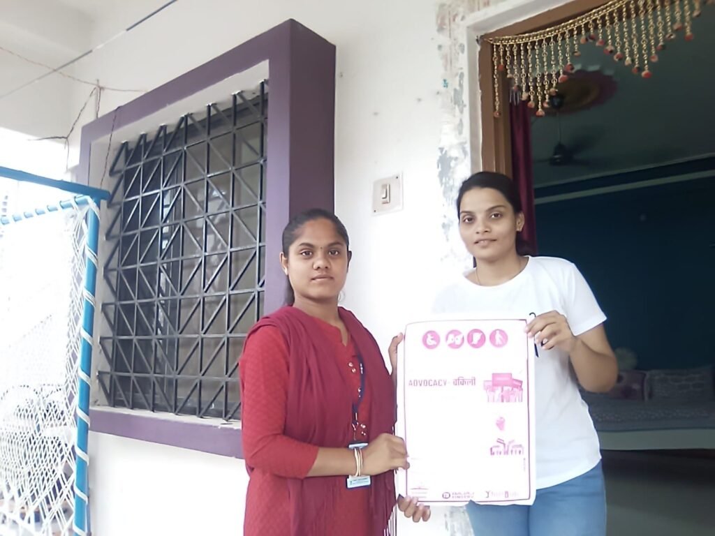 Nikitha and another woman holding a pink and white advocacy poster in Hindi, standing outside a building
