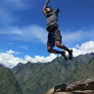 A person jumps joyfully off a rock with mountains and a bright blue sky in the background.