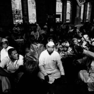A black-and-white photo of a group of people sitting closely together indoors, with one man in the center wearing a traditional cap and looking forward.