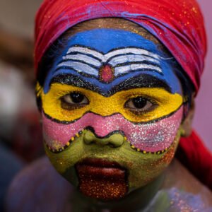 A child with vibrant, colorful face paint and a red headscarf looks directly at the camera.
