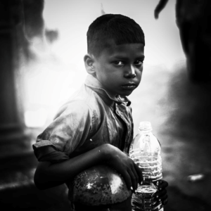 A young boy stands with a large water bottle in his hand, leaning against a metal post and looking directly at the camera.