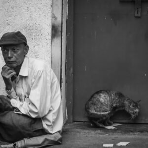 A man sits against a textured wall, resting his chin on his hand, while a cat crouches near a closed door beside him.