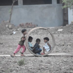 Four children play outdoors, with two sitting inside a large circular ring while another child pushes it across the ground.
