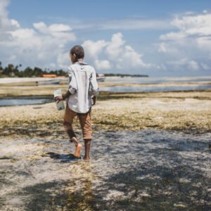 A person walking barefoot along a shallow, sandy shoreline under a bright sky with scattered clouds.