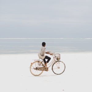 A person riding a bicycle along a wide, white sandy beach, looking out toward the calm, pale blue ocean under an overcast sky.