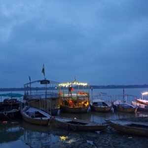 Boats anchored along a riverbank at dusk, with a brightly lit boat in the center reflecting light on the water under a cloudy blue sky.