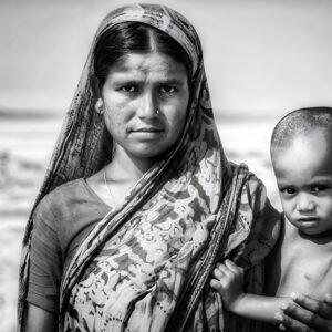 A black-and-white portrait of a woman wearing a sari standing outdoors, holding a young child close to her side.