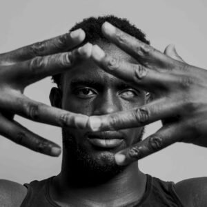 A black-and-white portrait of a man looking through his fingers, which are spread open in front of his face.