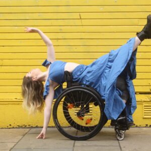 A woman in a blue dress performing a graceful dance pose in a wheelchair against a bright yellow wall.