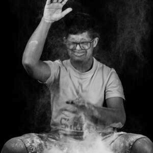A black-and-white photo of a man smiling while playing with powder or dust, which is rising around him
