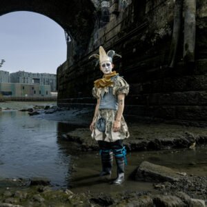 A person dressed in an elaborate clown costume and mask stands under a dark stone bridge near shallow water, with buildings visible in the background.