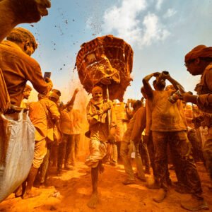 A vibrant scene of people covered in orange powder celebrating outdoors, with one person carrying a large basket on their head amid the crowd.