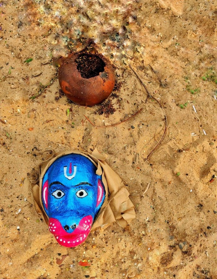 A vibrant hand-painted blue monkey mask with expressive features rests on a sandy surface, wrapped in cloth, evoking a cultural or spiritual essence. Nearby lies a broken earthen pot, partially filled with soil, adding a rustic and ritualistic touch to the scene, suggesting ties to traditional practices or ceremonies.