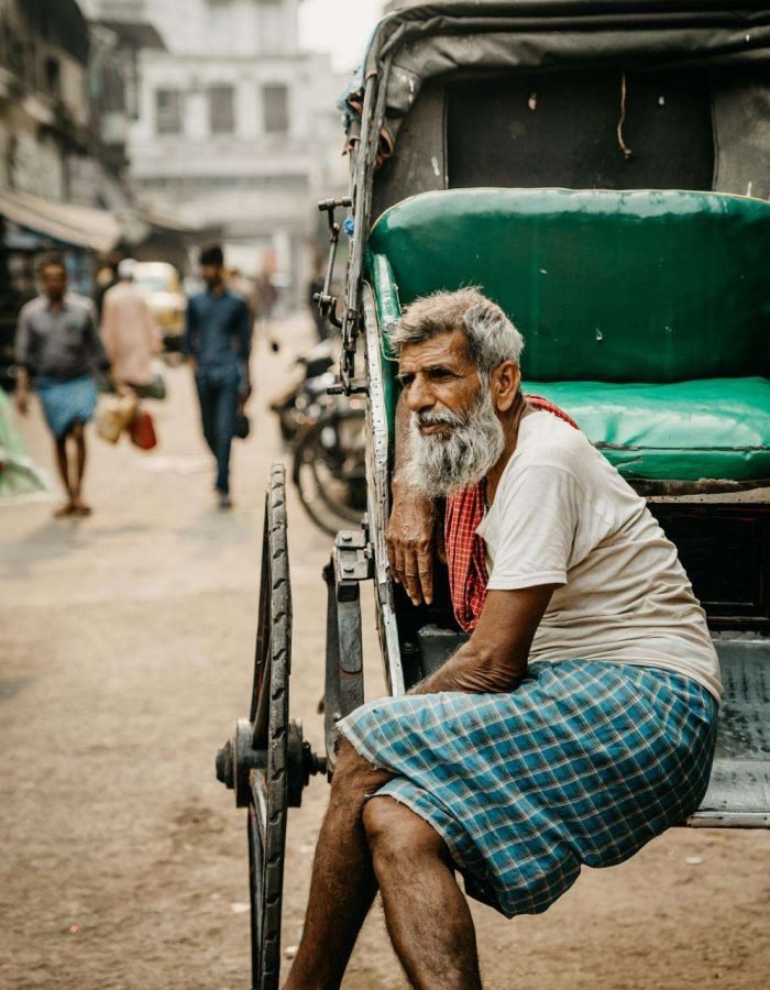 An elderly rickshaw puller with a gray beard and a contemplative expression sits on the edge of his rickshaw in a busy urban street. He wears a white shirt, a red towel draped around his neck, and a blue checkered lungi. The vibrant green seat of the rickshaw contrasts with the muted tones of the bustling street in the background, where pedestrians and vehicles move through the hazy atmosphere, capturing a moment of stillness amidst city life